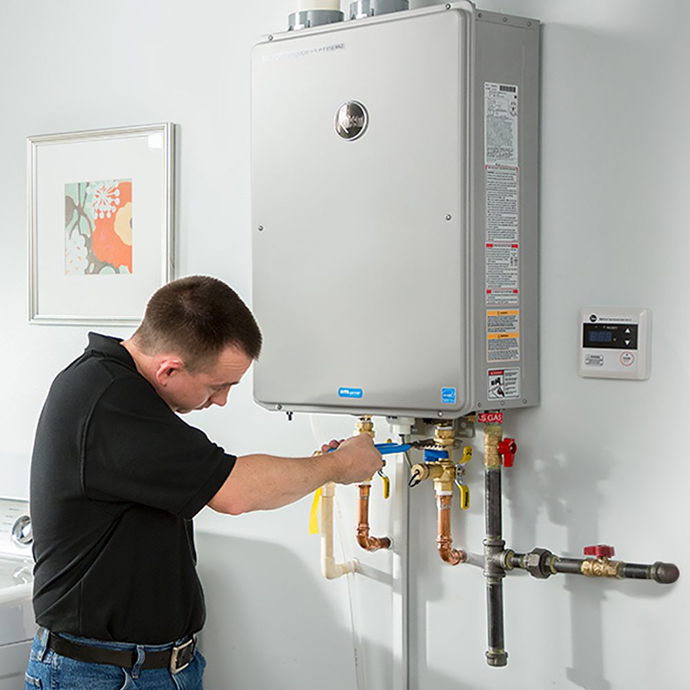 A technician in a black shirt working on the plumbing connections of a large, wall-mounted tankless water heater in a utility room.