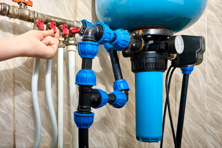 A hand adjusting a red valve on a complex plumbing manifold with a blue water filter and a large blue pressure tank visible.