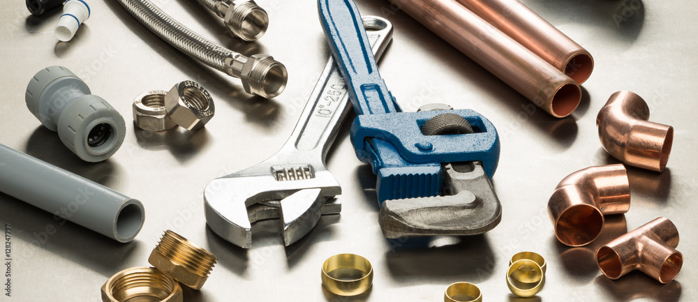 A collection of plumbing tools and materials, including an adjustable wrench, a pipe wrench, flexible hoses, copper pipes and fittings, and various connectors, laid out on a metallic surface.