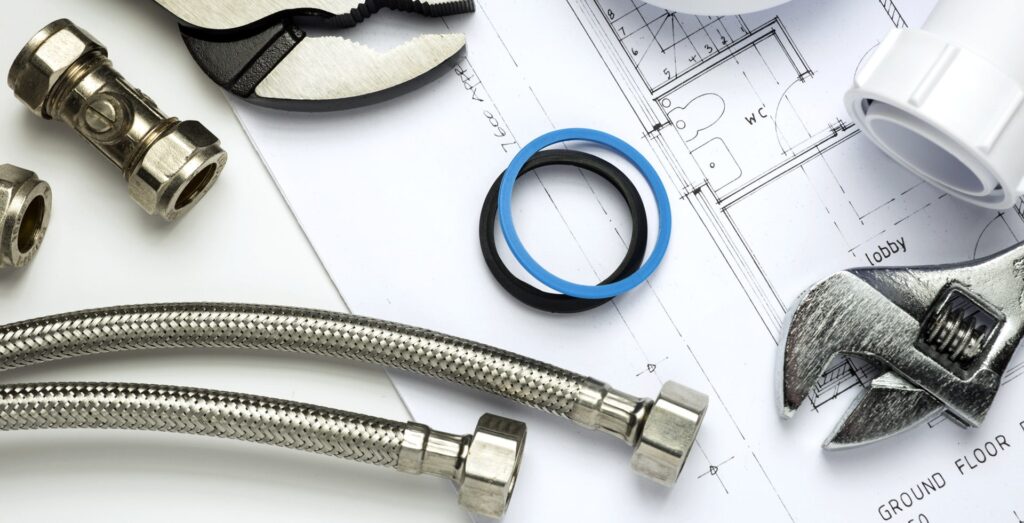 Various plumbing tools and components, including two braided stainless steel hoses, an adjustable wrench, pipe wrench, fittings, and O-rings, are laid out on top of a blueprint or architectural drawing with room labels like "WC" and "Lobby."