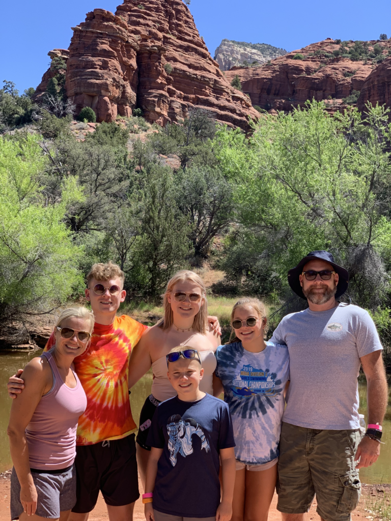 A family of six poses happily outdoors in a scenic rocky, mountainous landscape with lush green trees.
