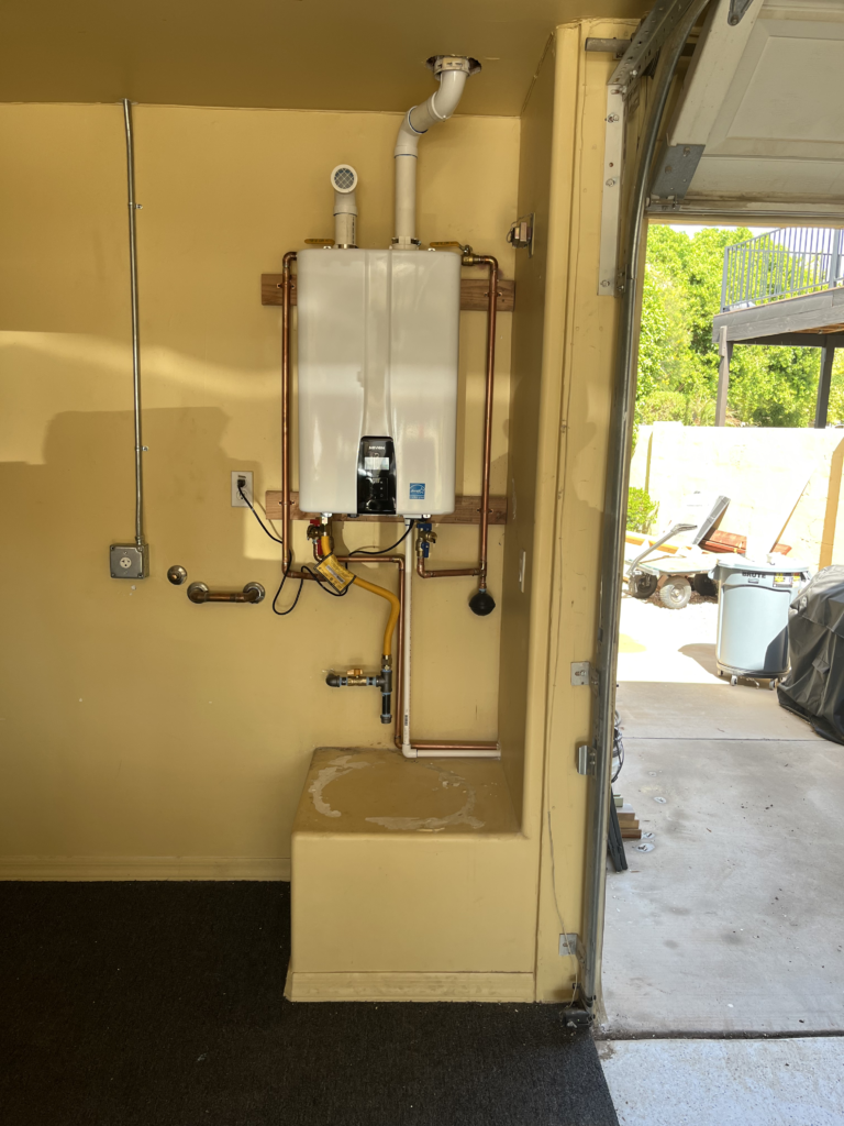 A white, rectangular tankless water heater is mounted on a yellow wall in a garage, with copper and PVC pipes connecting to it, and an open garage door reveals an outdoor view.