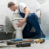 A male plumber in blue overalls and a white t-shirt leans over to fix a toilet in a grey tiled bathroom. A grey toolbox sits open to his left, and various hand tools are scattered on the floor in front of him.