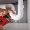 A plumber's hands hold a leaking white PVC pipe under a sink, with water dripping, and a red pipe wrench held nearby.