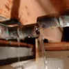A close-up of a burst and leaking copper pipe in a basement or crawl space, with water actively gushing and dripping onto the surrounding area.